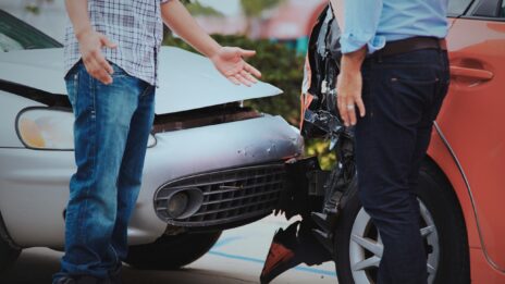 Two individuals visible from the waist down appear to negotiate after a fender bender, which is seen in the background,