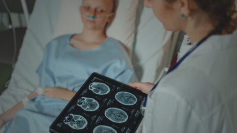 An over-the-shoulder view of a brain scan chart being shown to a young patient in a hospital bed.