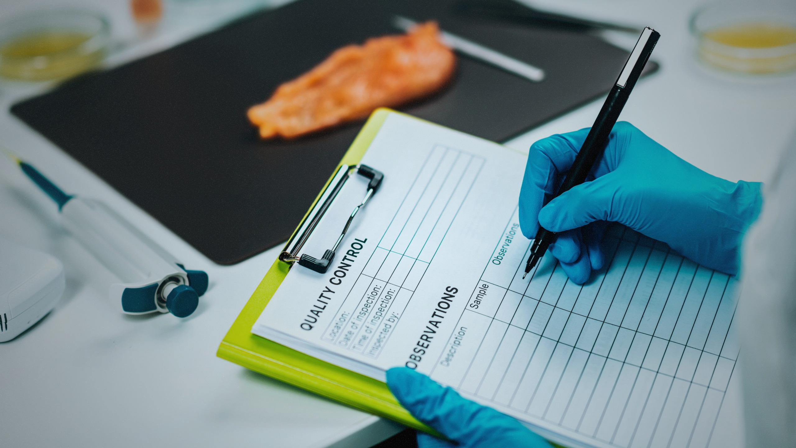 A gloved hand writes on a quality control clipboard in a lab setting.