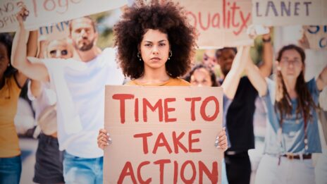 A woman holding a sign that reads 'Time to Take Action' stands in front of a protest, facing the camera.