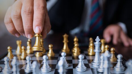 A close up of a chess board from the the perspective of one player. The opponent's hands are lifting a golden piece toward the foreground.